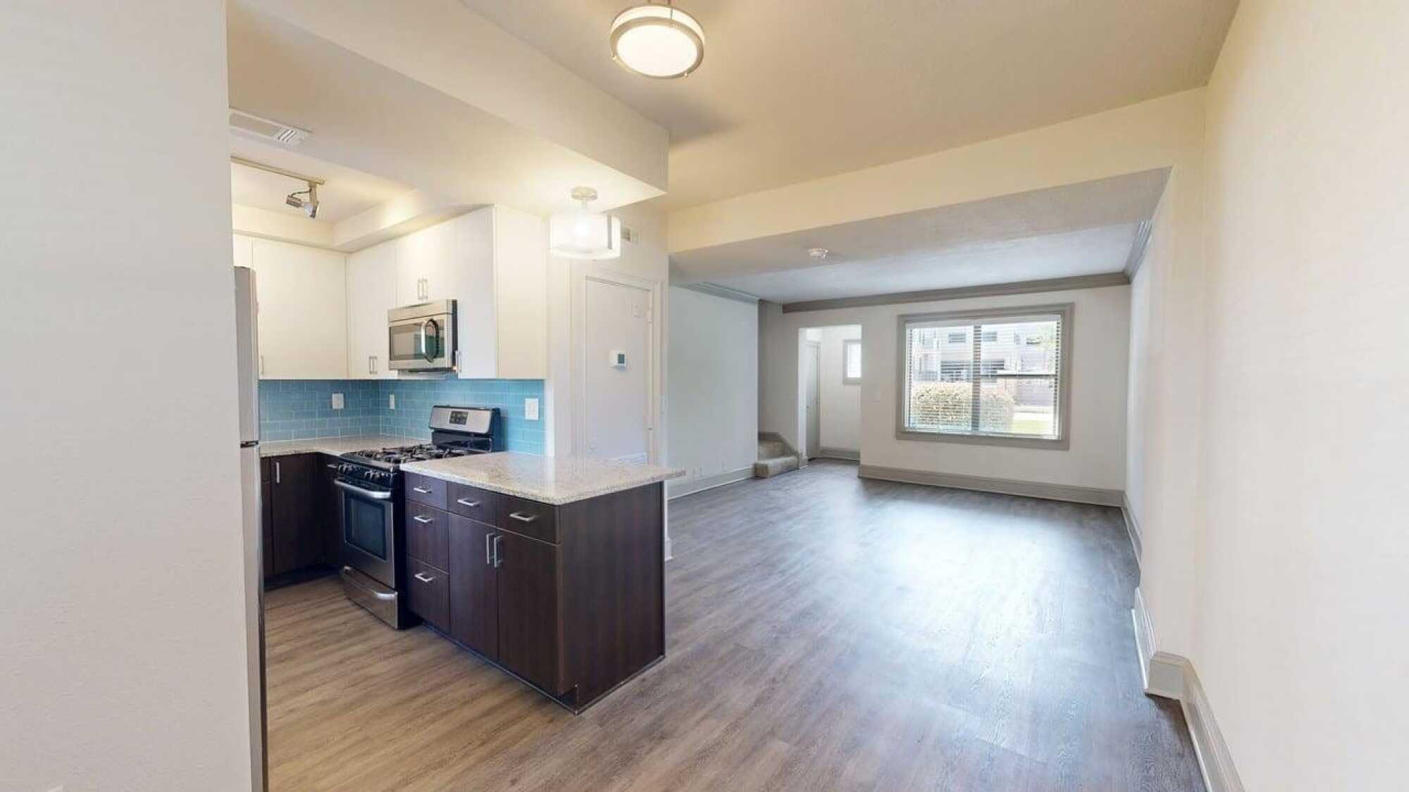 Modern apartment with an open kitchen, stainless steel appliances, wood floors, and large window in the living area.