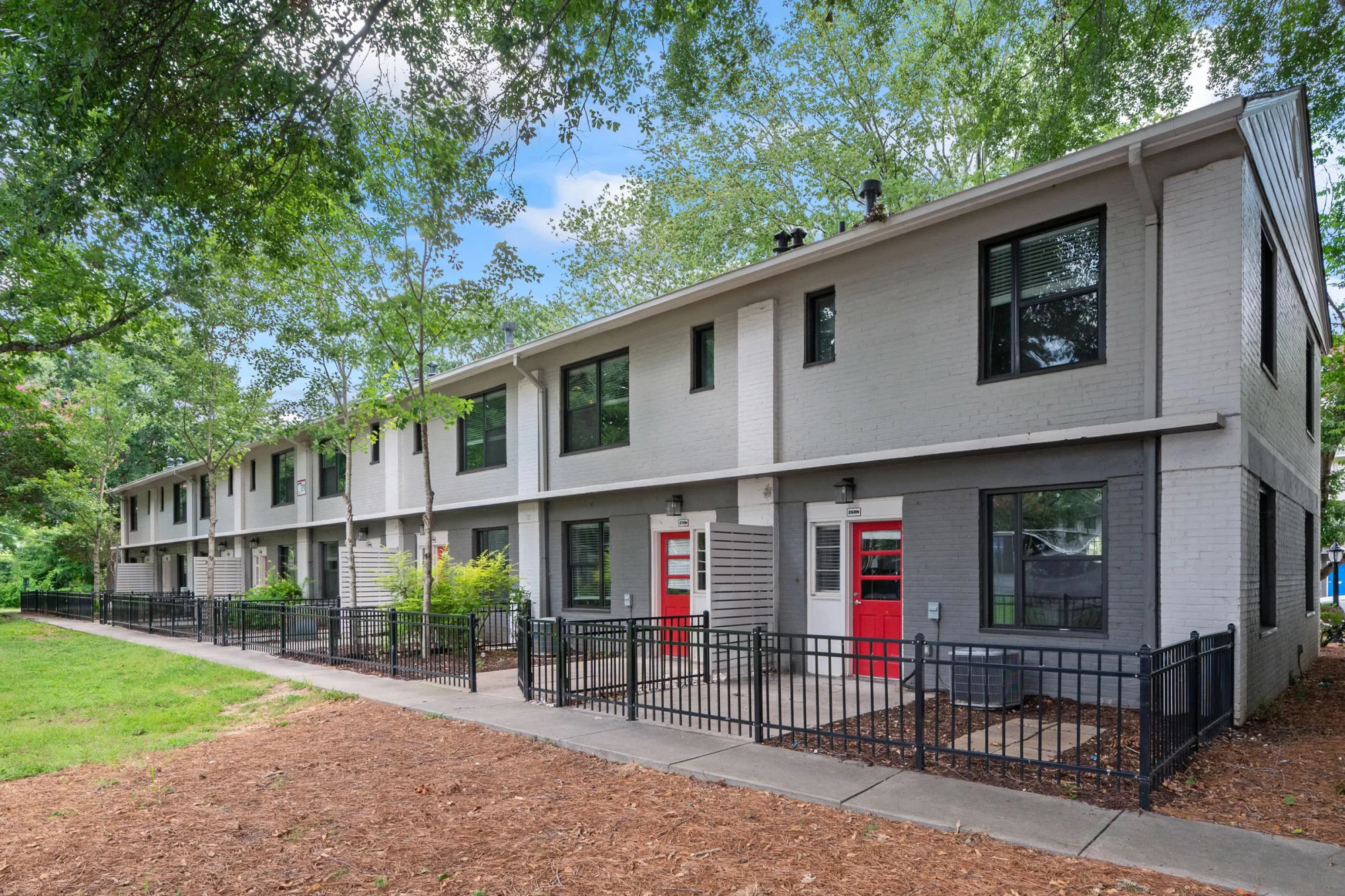 Updated two-story gray apartment building with red front doors, black wrought-iron fenced patios, mature trees, and a wood-mulched walkway leading along the building.