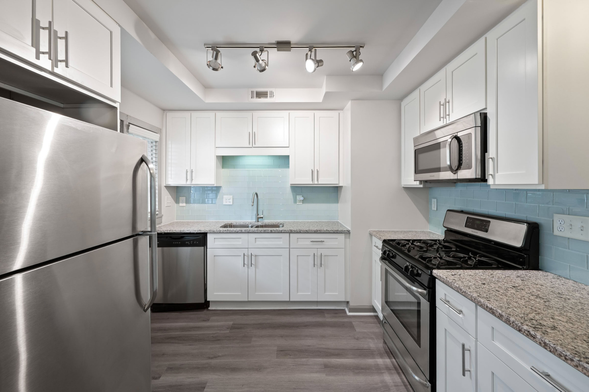Galley kitchen featuring crisp white cabinetry, light blue subway tile backsplash, granite counters, stainless steel refrigerator, dishwasher, microwave, and gas stove with gray wood-look flooring.