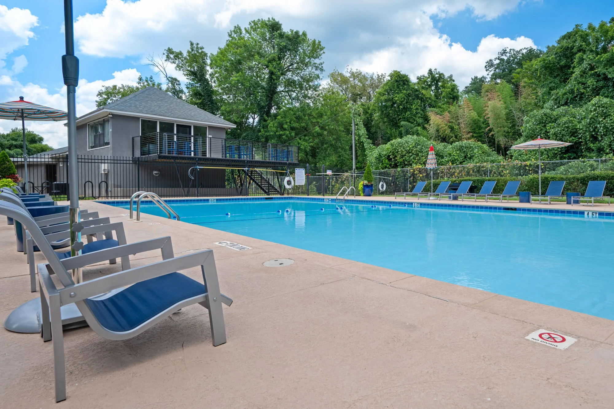 Inviting community pool with crystal blue water, lounge chairs lined along paver decking, vibrant umbrellas, and a contemporary gray pool house surrounded by trees.